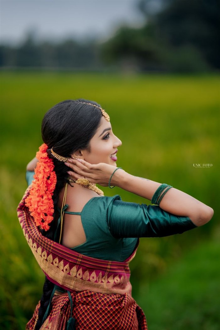 Bridal side profile in green saree with orange floral hair garland, paddy field backdrop, Jaffna bridal makeup