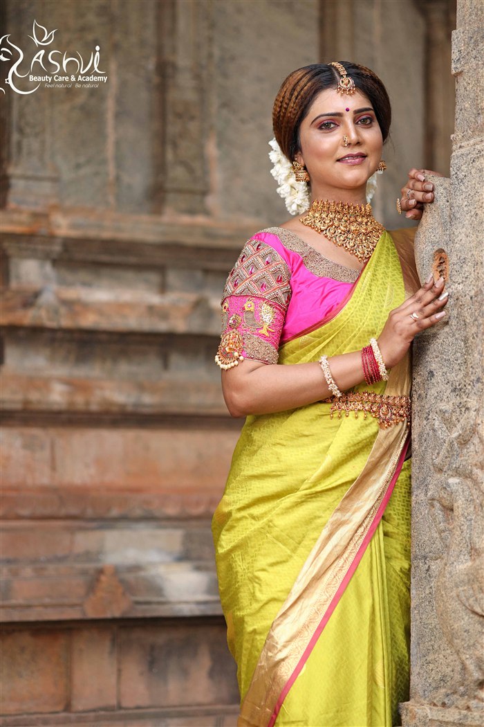 Bride in yellow silk saree with gold jewellery leaning against a heritage temple pillar, Ashvi Beauty Care Jaffna