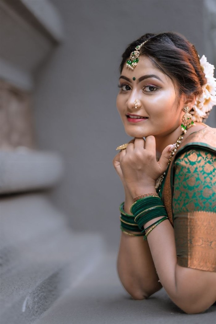Bride in green and gold Kanjivaram silk saree with emerald jewellery, leaning forward on textured stone steps, Ashvi Beauty Care Jaffna